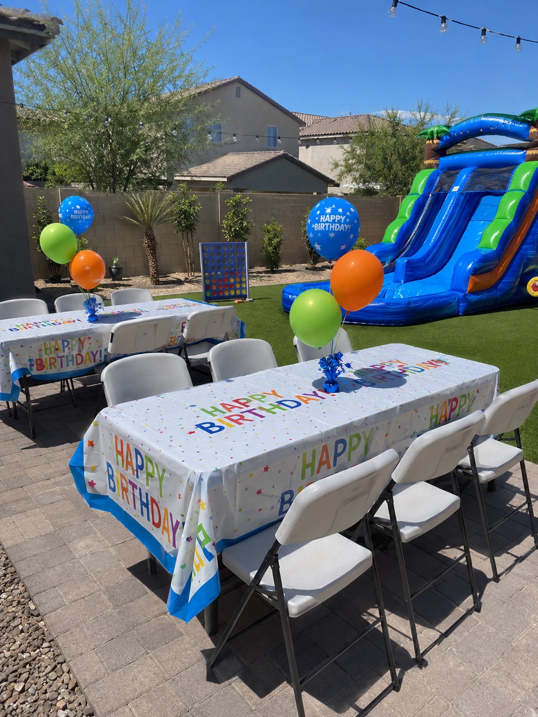 Backyard birthday party setup with tables, white folding chairs, balloons, giant Connect 4, and an inflatable slide